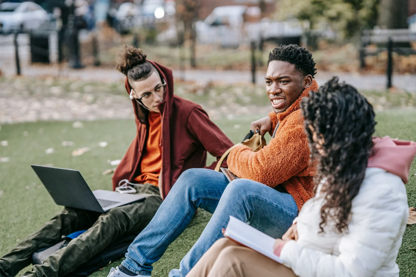 Students sit on the grass and study together using a laptop and book.
