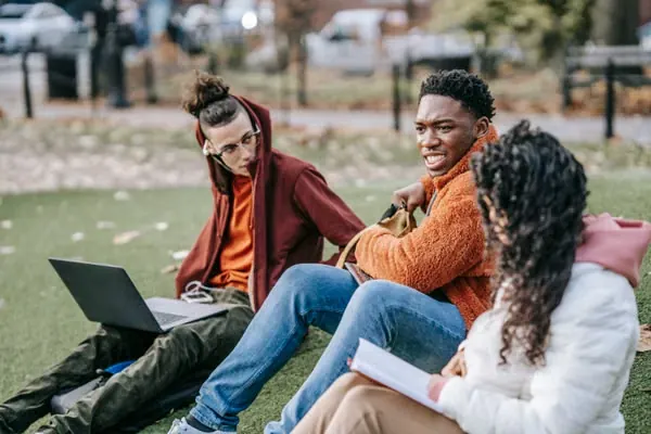 Group of multiracial students sitting on grassy ground with book browsing laptop while studying together on street against blurred background