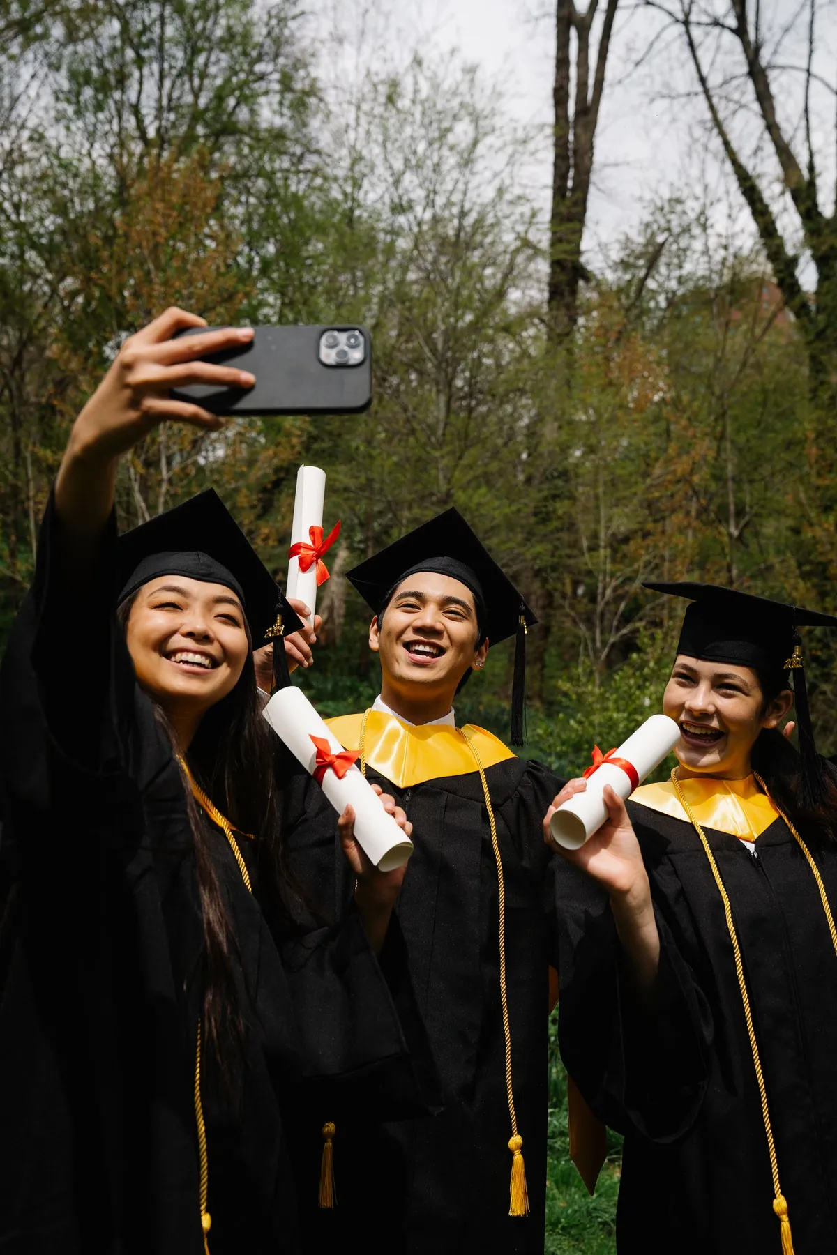 Three happy graduates in caps and gowns take a selfie with diplomas outdoors.